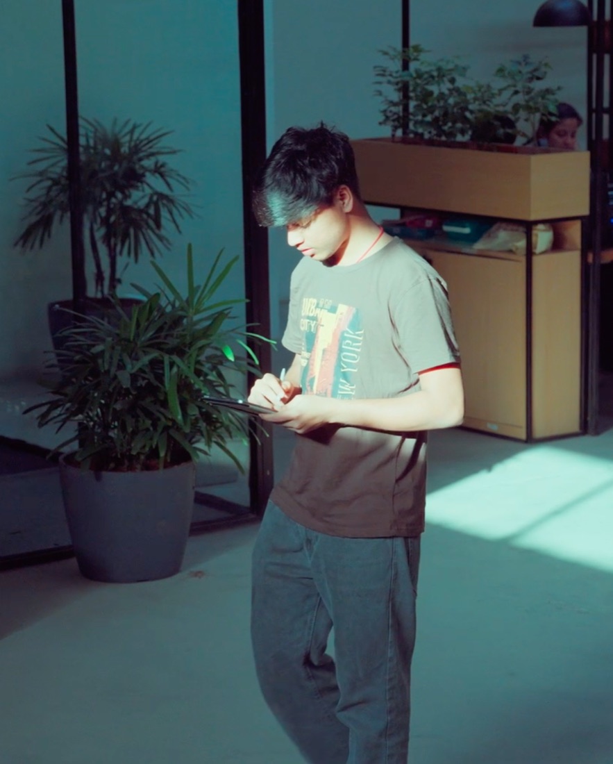Pavan writing on a tablet beside potted plants, with sunlight on the floor behind him